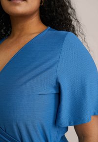 Royal blue zavinovací top s krátkými volánovými rukávy. Texturovaná tkanina má jemný vzor, který zdůrazňuje výstřih do V a nařasený pas.