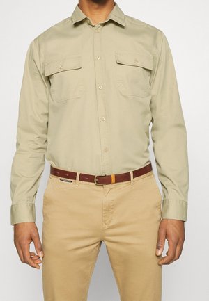 Homme portant une chemise beige à manches longues avec deux poches poitrine et un pantalon beige avec une ceinture marron.