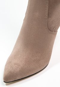 Bota corta de ante beige con punta afilada y textura suave. Cuenta con un panel elástico lateral para facilitar el calzado. Diseño de un solo color, sin herrajes.