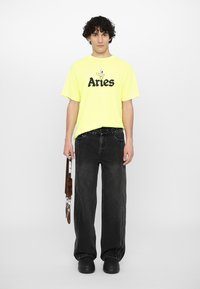 T-shirt in cotone giallo neon con la scritta "Aries" stampata in nero, abbinata a jeans in denim nero dal taglio loose fit e scarpe con plateau nere.