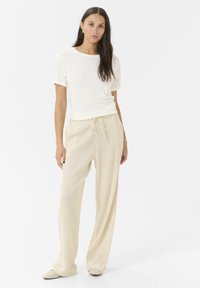 T-shirt blanc léger associé à un pantalon beige ample à cordon de serrage avec un motif texturé. Le modèle se tient devant un fond uni.
