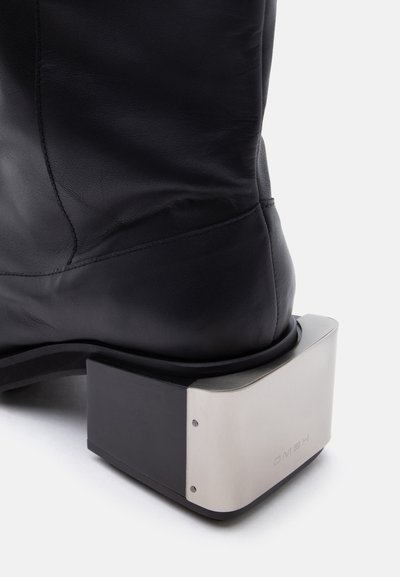 Μαύρο δερμάτινο μποτάκι με παχιά, τετράγωνη μεταλλική φιάπη σε βουρτσισμένο ασημί φινίρισμα και ορατές βίδες.