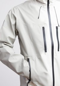 Giacca impermeabile grigio chiaro con cappuccio, due tasche frontali con zip e dettagli zip neri. Texture liscia e design minimale.