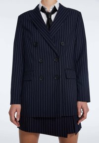 Laivastonsininen kaksirivinen bleiseri, jossa valkoiset raitakuosit, kuusi mustaa nappia, läppätaskut ja jäsennelty istuvuus. Yhdistetty minihameeseen.