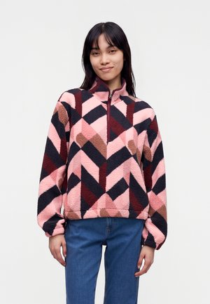 Femme portant un pull polaire multicolore avec des motifs géométriques et un col à demi-zip, associé à un jean bleu.