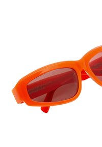 Occhiali da sole in plastica arancione con bordi smussati, lenti di un rosso scuro, e iscrizione del logo sul lato interno della montatura. Design retrò.
