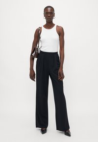 Schwarze weite Beinhosen mit Falten, kombiniert mit einem weißen ärmellosen Oberteil. Das Outfit wird durch eine kleine braune Tasche und spitze schwarze Schuhe ergänzt.