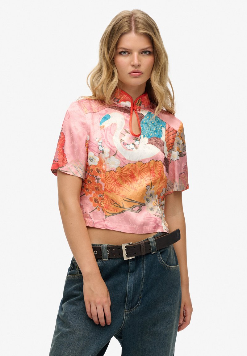 Blusa di seta rosa tagliata con motivi floreali, maniche corte e colletto alla mandarina. Indossata con jeans denim a vita alta e una cintura marrone.