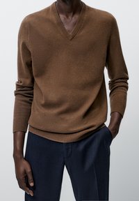 Homme portant un pull marron à col en V à manches longues et un pantalon bleu foncé, une main dans la poche, debout devant un fond uni.