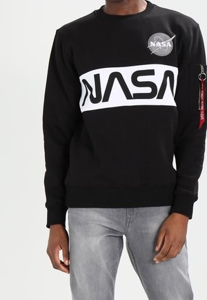 Person iført sort NASA sweatshirt med hvidt logo bånd tværs over brystet og patches på øverste venstre og venstre ærme, kombineret med lysegrå jeans.