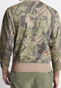 Felpa camouflage in verde, marrone e beige con un motivo di corteccia d'albero e foglie. Polsini e orlo a coste in un morbido tessuto color sabbia.