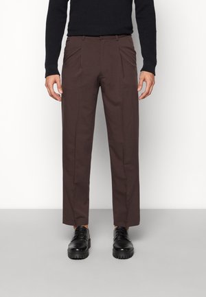 Mand iført mørkebrune bukser med straight-fit, sorte snørebåndssko og en sort, langærmet tekstureret sweater, stående mod en ensfarvet baggrund.