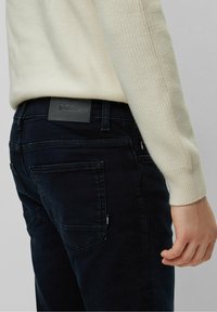 Dark blue denim jeans with a smooth texture, featuring a black leather logo patch on the back waistband and multiple pockets.