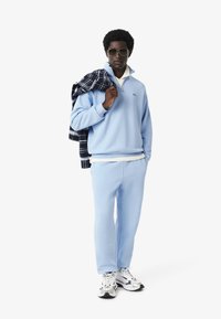 Sudadera de forro polar azul claro con media cremallera, pantalones jogger a juego y camiseta blanca. El modelo lleva gafas de sol negras y una chaqueta de cuadros.