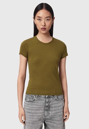Kvinde med glat, mørkt hår har en olivengrøn kortærmet t-shirt og grå jeans på og står mod en ensfarvet lys baggrund.