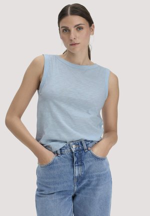 Vrouw met een mouwloos lichtblauw topje en een blauwe high-waisted jeans, met handen in de zakken, staande tegen een effen achtergrond.
