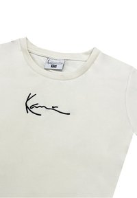 Krämfärgad t-shirt med kort ärm, tillverkad av mjukt tyg och med en svart broderad signatur på framsidan. Halsringningen är rund. Storlek: XS.