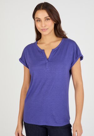 Femme portant un t-shirt henley violet à manches courtes retroussées, debout devant un fond clair uni, souriant légèrement.