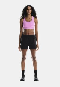 Sostén deportivo rosa con tirantes anchos, combinado con pantalones cortos deportivos negros. La modelo lleva calcetines y zapatos negros, de pie contra un fondo blanco.
