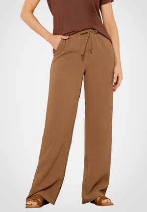 Frau trägt hoch taillierte, lockere braune Hose mit Kordelzug in der Taille und Taschen, kombiniert mit braunen Sandalen und einem braunen Oberteil.