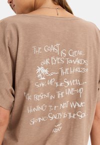 Hnědé tričko s krátkým rukávem, které na zadní straně obsahuje bílý text a grafiku palmového stromu. Měkká textura, volný střih, casual design.