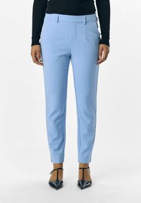 Světle modré kalhoty na míru z hladké tkaniny, s předními sklady a slim fit střihem. Spárováno s černými lodičkami na kotníkovém pásku.