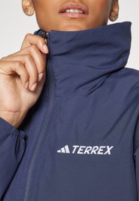 Jachetă bleumarin cu fermoar, fabricată din material ușor, cu guler ajustabil și logo-ul brodat "TERREX" în alb.