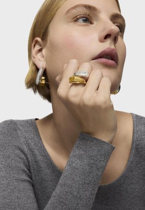 Mujer con cabello corto y rubio, lleva grandes pendientes de aro grises y anillos gruesos de plata y oro, apoyando la barbilla en la mano, con una blusa gris.