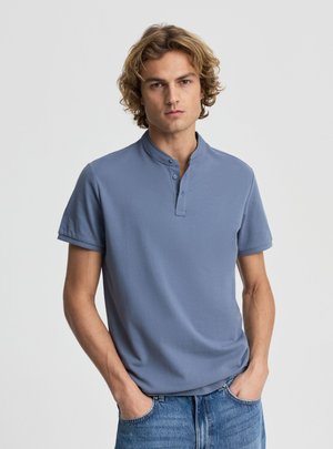 Giovane uomo con capelli biondi ricci che indossa una maglietta henley blu a maniche corte e jeans blu, mani nelle tasche, espressione neutra.