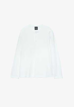 Bijela košulja dugih rukava, mekani materijal, okrugli izrez, plaket s tri gumba, ušiveni križni detalj na prsima, opušteni kroj, minimalistički dizajn.