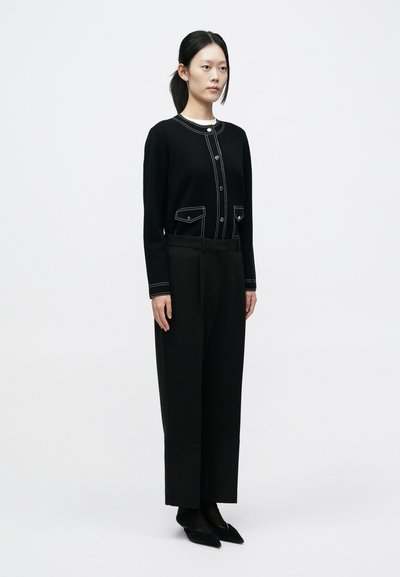 Fekete kötött kardigán kontrasztos fehér varrással, elülső zsebekkel és gombokkal, öltönyre szabott fekete nadrággal és hegyes orrú cipővel párosítva.