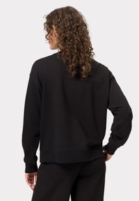 Černá mikina vyrobená z měkké tkaniny, s volným střihem, dlouhými rukávy, žebrovanými manžetami a rovným lemem. Zadní pohled ukazuje kudrnaté vlasy a uvolněný střih.