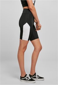 Pantalones cortos deportivos negros con un panel lateral blanco, hechos de material elástico; de longitud hasta la mitad del muslo; combinados con zapatillas negras y blancas.