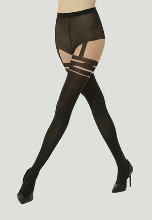 Onderlichaam met zwarte gedessineerde panty's en zwarte hoge hakken, benen gekruist op midden dijhoogte tegen een lichtgrijze achtergrond.