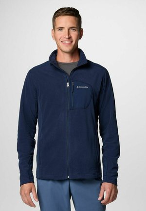 Marineblaue Fleecejacke mit einer Reißverschlusstasche auf der Brust, hohem Kragen und langen Ärmeln. Sie hat eine glatte Textur und das Columbia-Logo auf der Tasche.