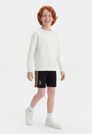 Niño pelirrojo sonriente que lleva una sudadera blanca, pantalones cortos deportivos negros con rayas doradas, calcetines blancos y zapatillas blancas, caminando hacia adelante.