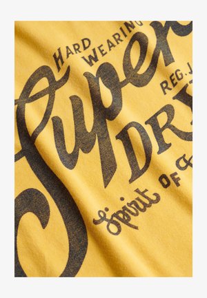 Tela amarilla con texto de estilo vintage en negro que dice "Superdry Hard Wearing Spirit of" y letras adicionales parcialmente visibles.