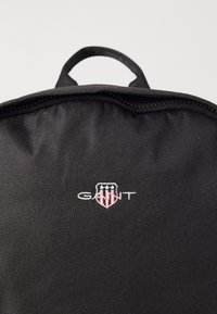 Sac à dos en tissu noir avec une poignée supérieure et une fermeture éclair. L'avant présente un logo brodé blanc avec un écusson et des accents rouges.