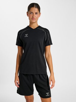 Ung kvinde iført sort hummel sportst-shirt og shorts, stående mod en ensfarvet hvid baggrund med neutralt udtryk.