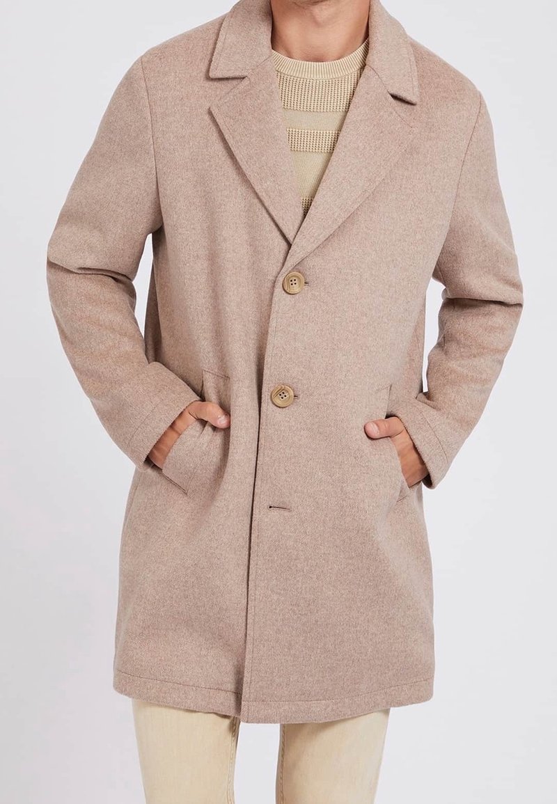 Beige uldfrakke med reverskrave og to knapper, båret over en struktureret beige sweater, hænder i frakkens lommer.