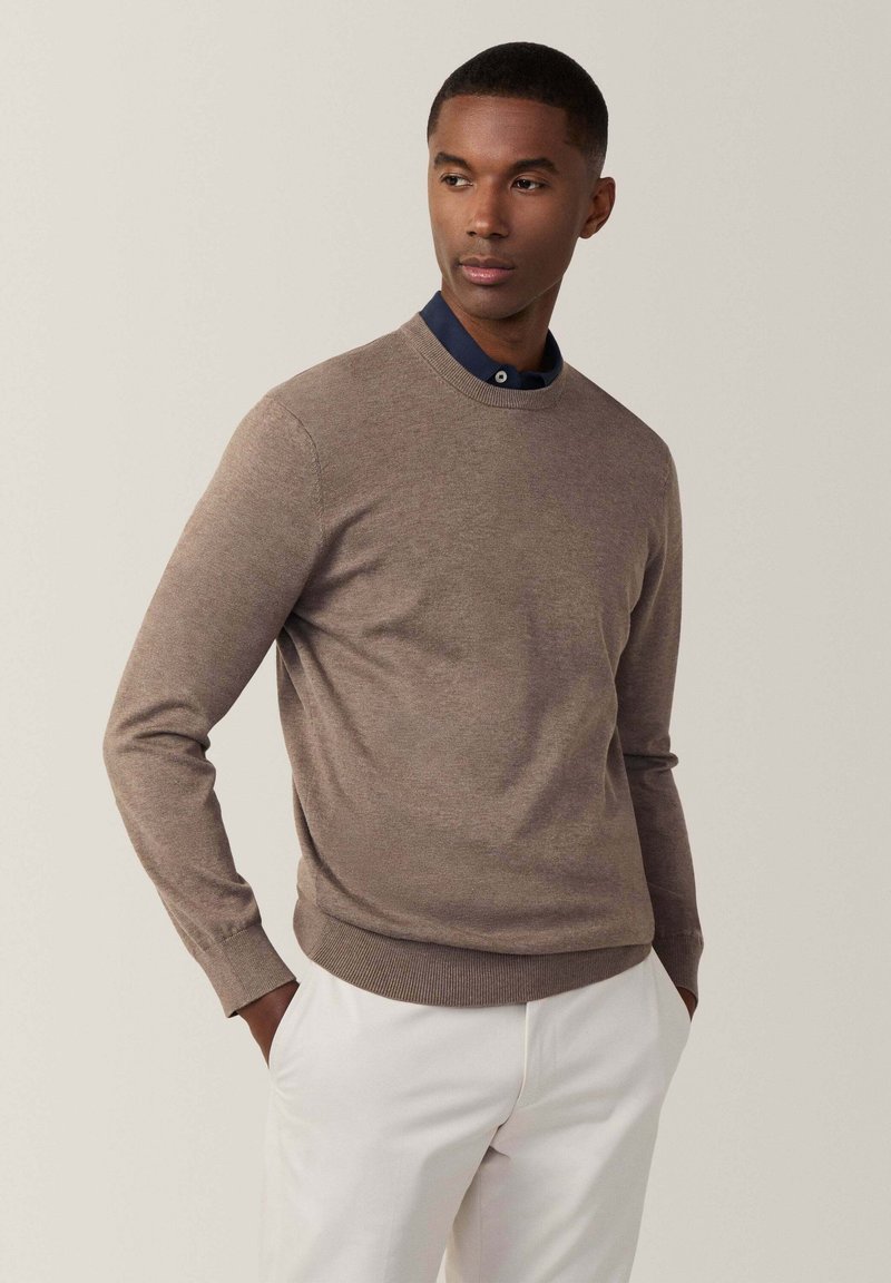 Braun gestrickter Pullover mit Rundhalsausschnitt und Rippensaum, getragen über einem navyblauen Hemd mit Kragen, kombiniert mit weißen Hosen.