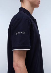 marinblå polo skjorta med krage, som har en vit detalj på ärmarna och broderi "Napapijri Geographic Since 1987" på armen.