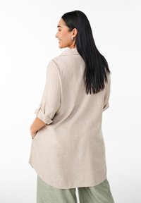 Camicia di lino beige chiaro con maniche arrotolate, colletto e orlo curvo, abbinata a pantaloni verde salvia. Tessuto liscio e vestibilità rilassata.