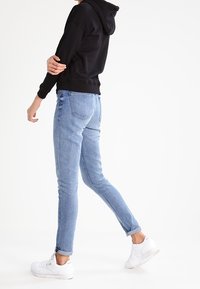 Jeans in denim blu aderenti con una leggera scoloritura, cinque tasche e orli arrotolati, abbinati a una felpa nera con cappuccio e sneakers bianche.