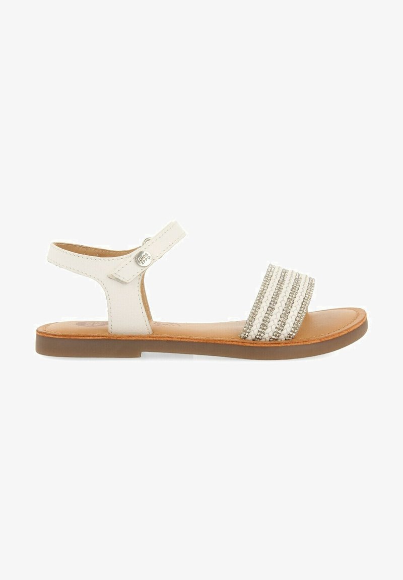 Sandalia de cuero blanca con suela plana, que presenta una tira delgada en el tobillo y un empeine ancho con adornos a rayas plateadas y blancas.