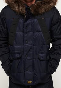 Giacca imbottita blu navy con cappuccio foderato in pelliccia sintetica, dettagli neri, bottoni a pressione anteriori e due tasche frontali. Presenta una texture trapuntata.