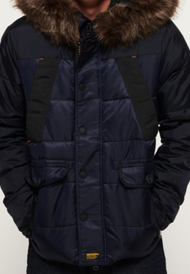 Giacca imbottita blu navy con cappuccio foderato in pelliccia sintetica, dettagli neri, bottoni a pressione anteriori e due tasche frontali. Presenta una texture trapuntata.