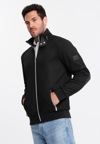 Giacca nera con zip e colletto alto, texture a coste, zip bianca a contrasto e tasche frontali. Presenta dettagli grigi sulle maniche.