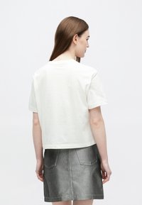 Žena s rovnými hnedými vlasmi, oblečená v bielej krátkym rukávom tričku a sivej rifľovej sukni, stojaca pred jednoduchým bielym pozadím.