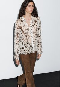 Blouse légère et fluide à base blanche avec des motifs abstraits marron. Associée à un pantalon évasé en suède marron, avec une coupe décontractée et une texture riche.